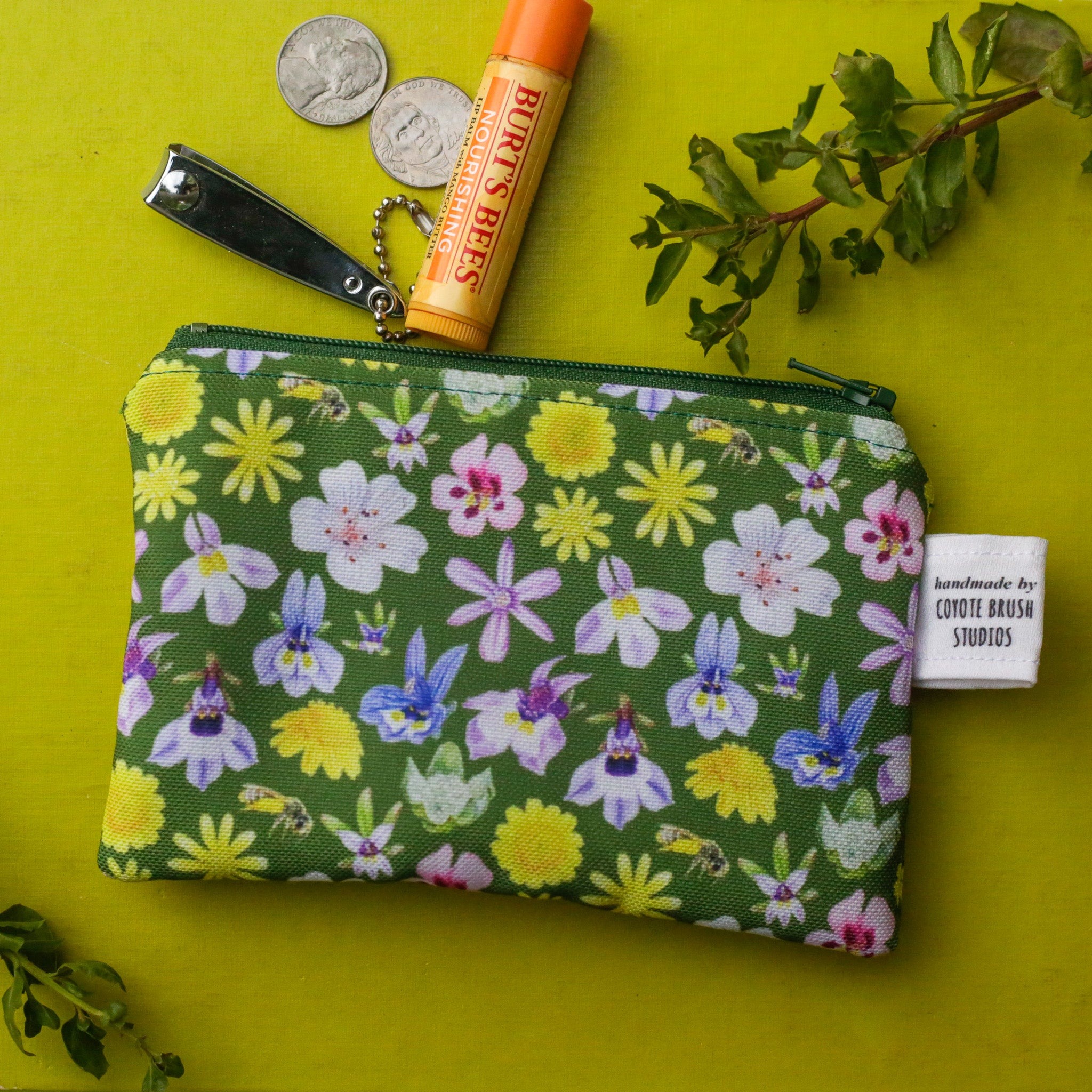 A green zipper pouch featuring vernal pools flowers on a bright green background next to coins, nail clippers, and lip balm. 