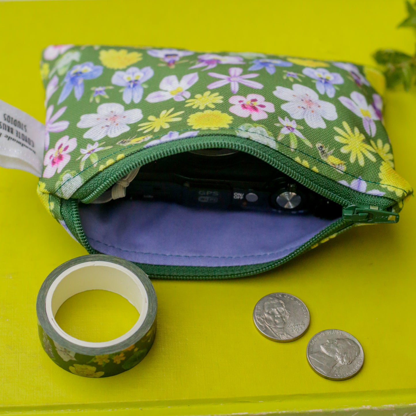 Camera is inside the pouch. Outside the pouch is a roll of washi tape and some coins. 
