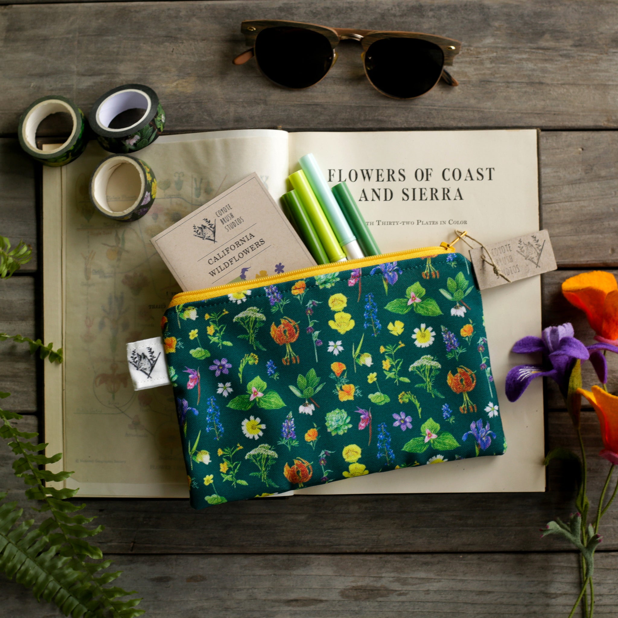 Native wildflowers printed on a dark green pouch with golden yellow zipper surrounded by stationery supplies. 
