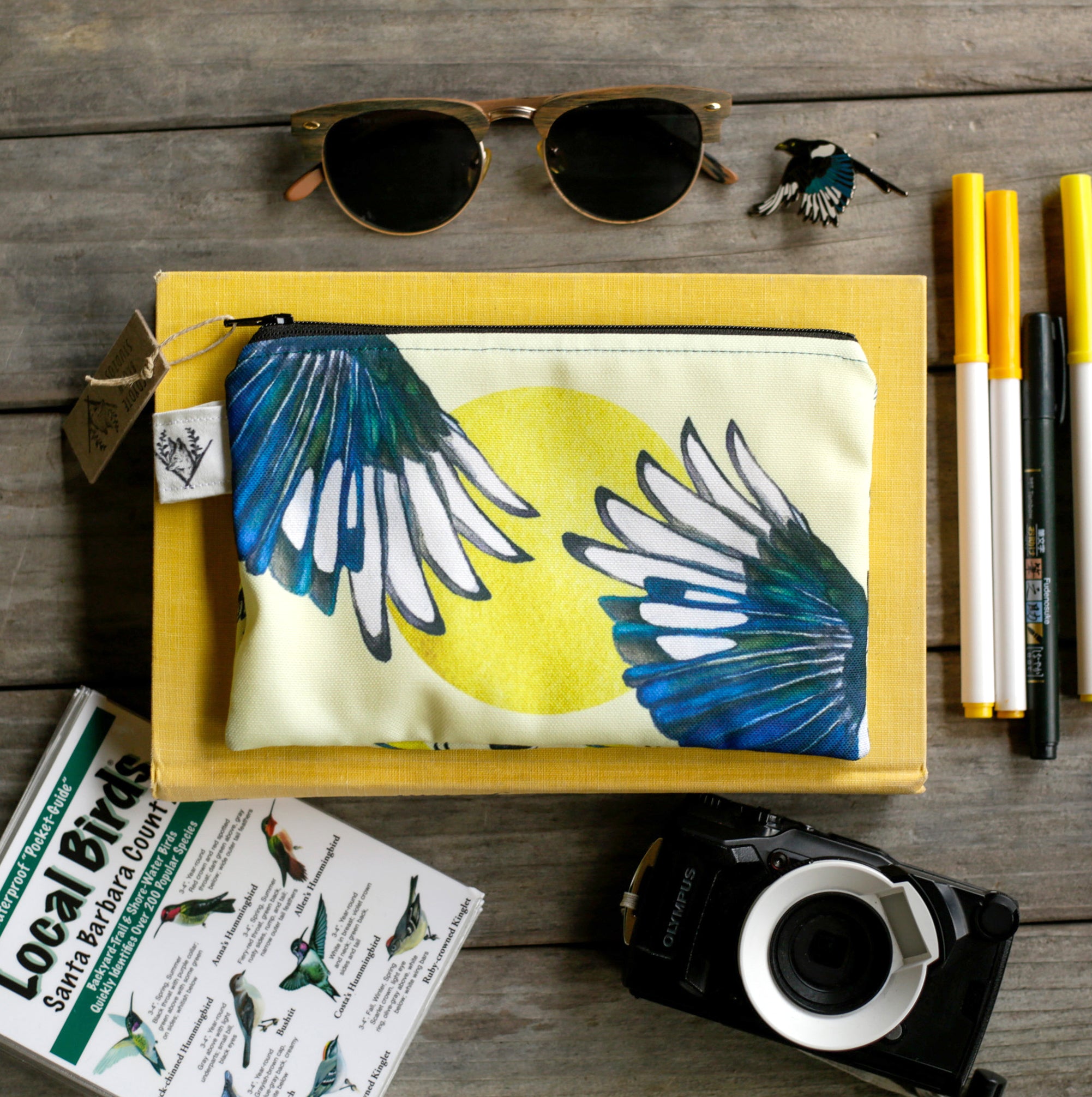 Yellow pouch with outstretched magpie wings surrounded by markers, sunglasses, a camera, and a bird guide. 