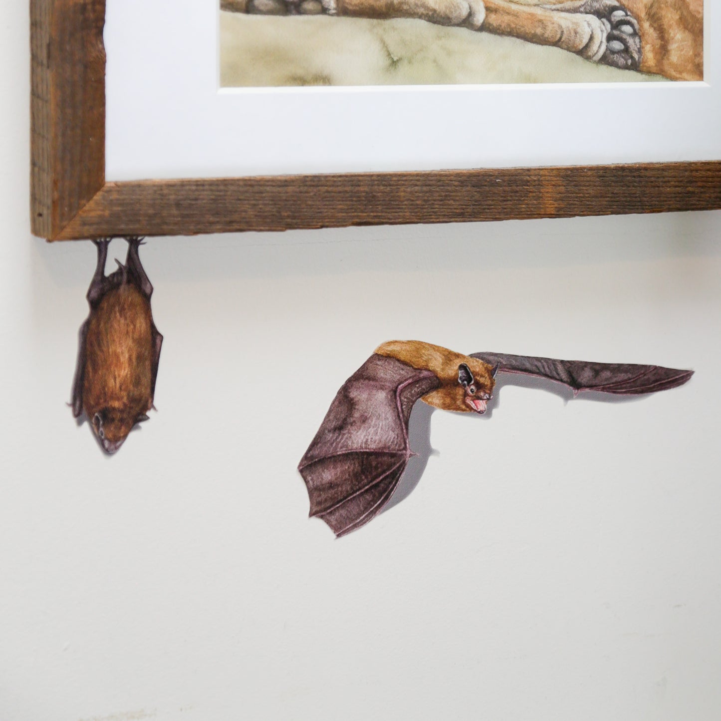 Realistic Big Brown Bat roosting underneath a picture frame next to another bat in flight.